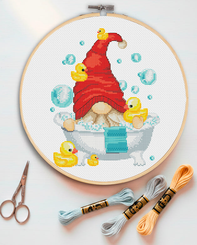 Embroidery hoop with a gnome and rubber duck design, surrounded by embroidery threads and scissors on a light background.