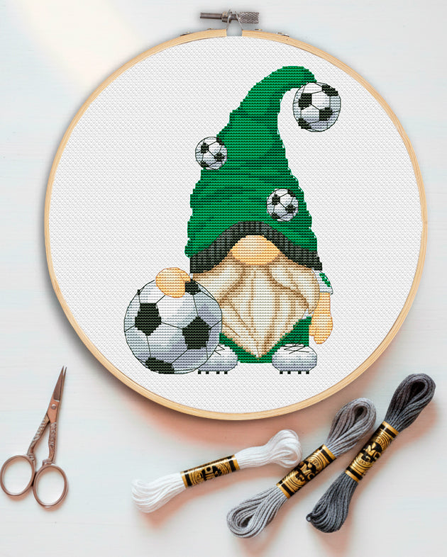 Cross-stitched gnome with soccer balls in an embroidery hoop, with scissors and thread on a light background.