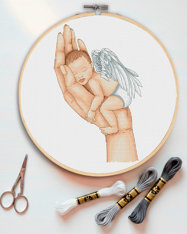 Embroidery hoop with a design of a hand holding a baby angel, accompanied by embroidery threads and scissors on a light background.