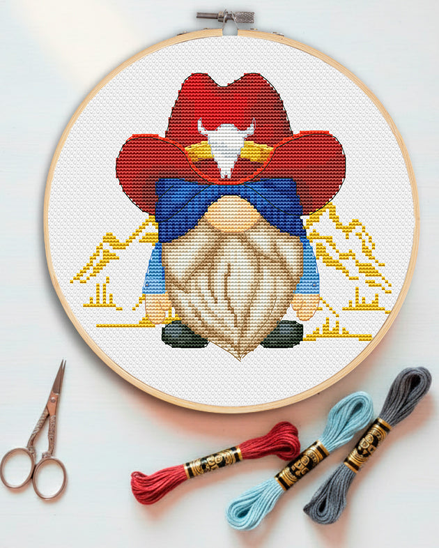 Wild West Gnome Cross Stitch Pattern. Embroidery hoop with a gnome design, cross-stitched on white fabric, with embroidery threads and scissors on a light background.