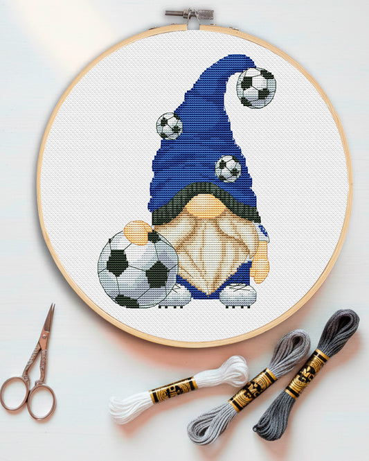 Embroidery hoop with a gnome holding soccer balls, surrounded by embroidery threads and scissors on a light background.