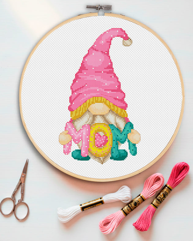 Cross-stitched gnome with a pink hat on a white background, framed in gold.
