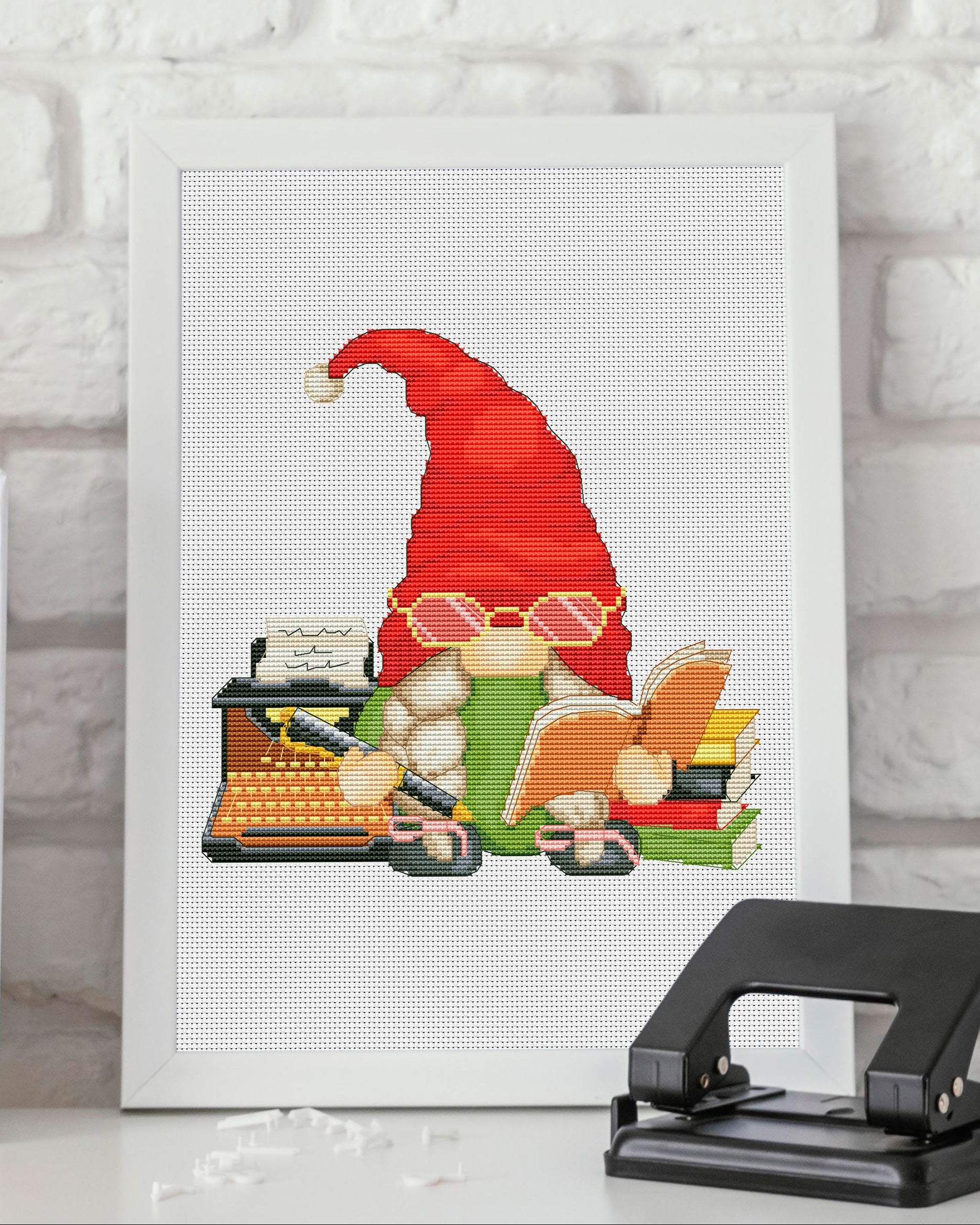 Cross-stitched gnome with a typewriter on a white background with books and a hole puncher.