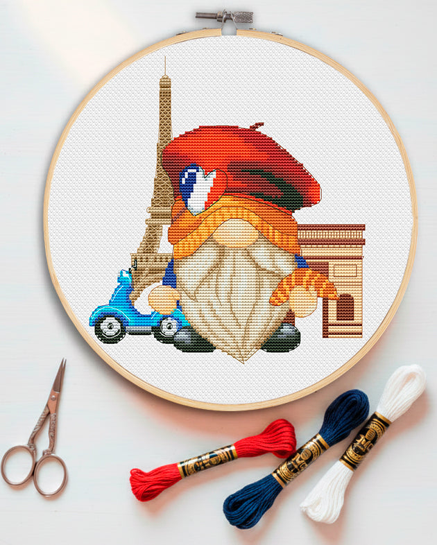 Cross-stitched gnome with a red beret and blue car in an embroidery hoop, with threads and scissors on a white background.
