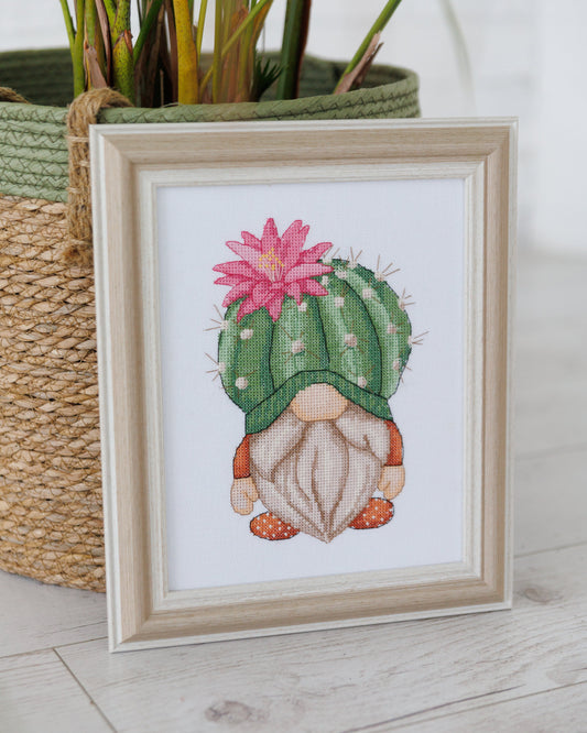 Framed cross-stitched picture of a cactus gnome with a pink flower, placed next to a woven basket.