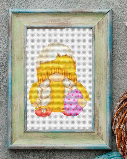 Cross-stitched gnome with a mushroom and Easter eggs in a wooden frame on a textured surface.