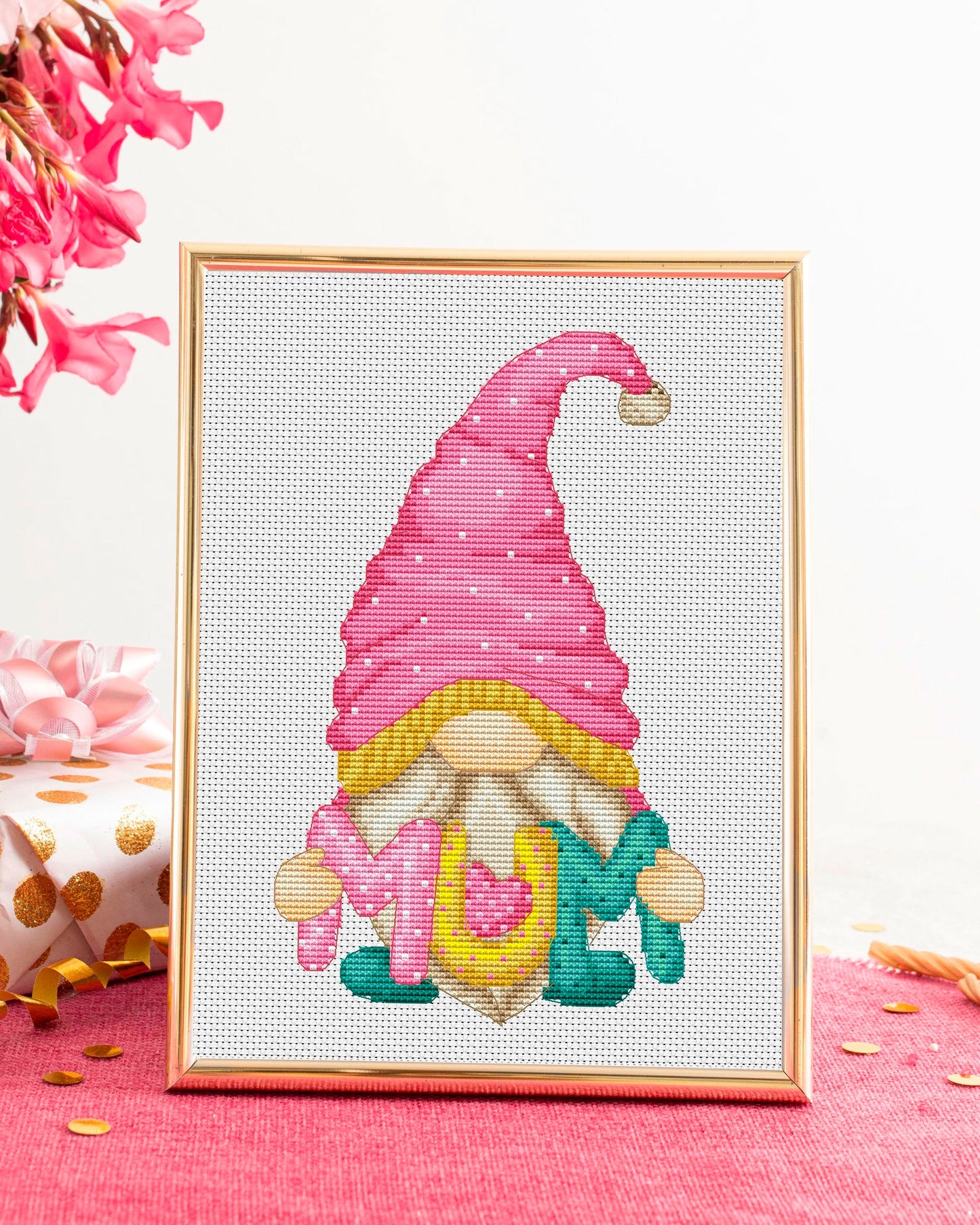 Cross-stitched gnome with a pink hat on a white background, framed in gold.