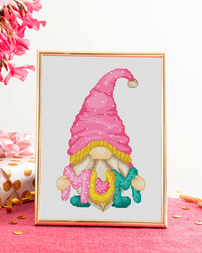 Cross-stitched gnome with a pink hat on a white background, framed in gold.