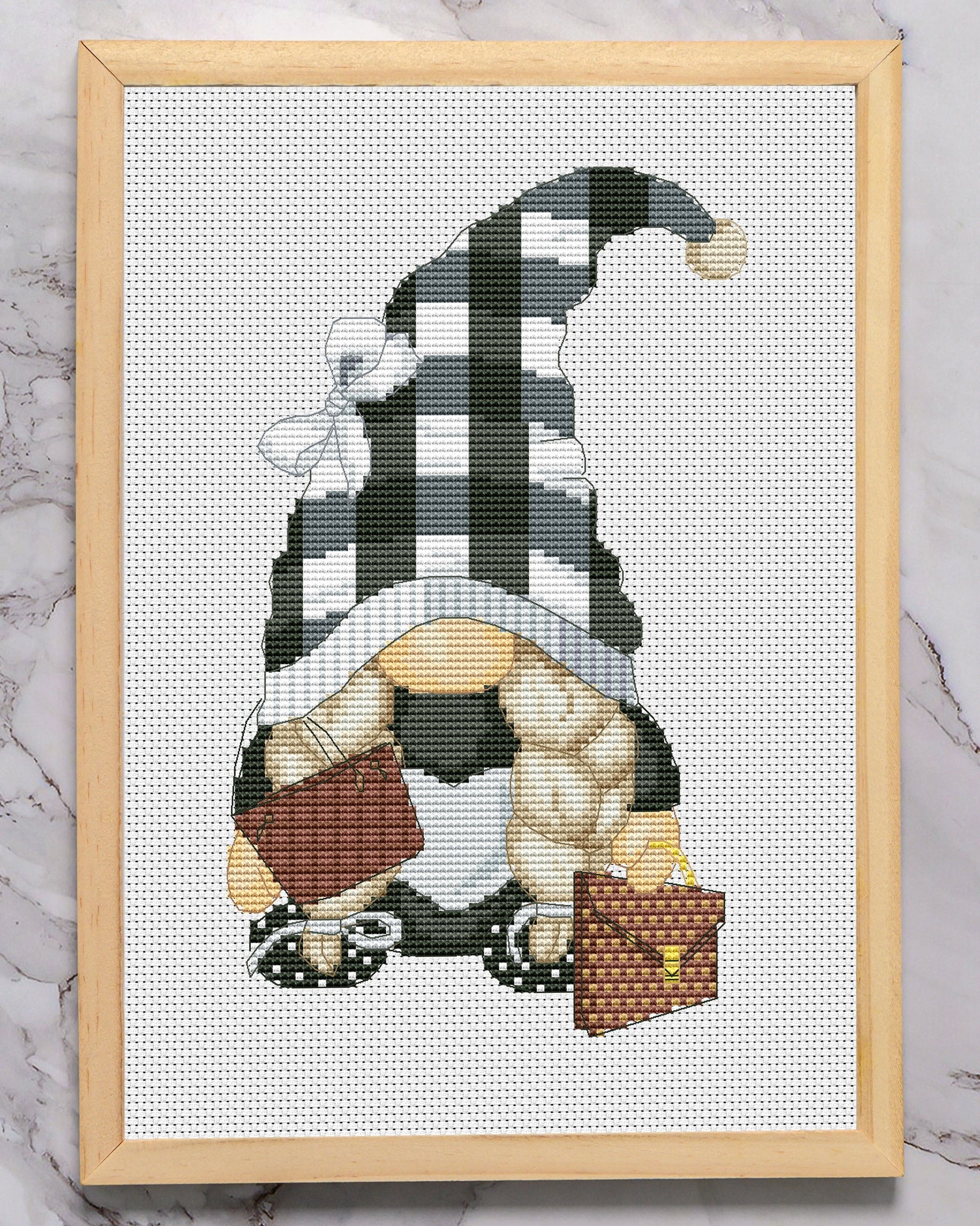 Cross-stitched gnome with a checkered hat in a wooden frame on a marble surface with glasses above.