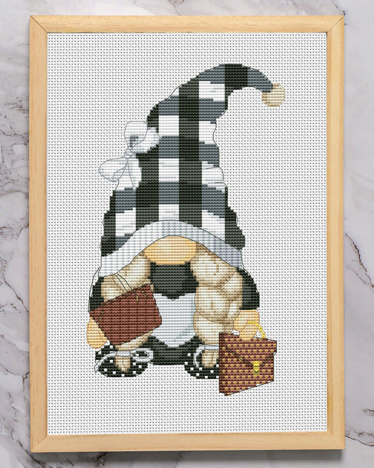 Cross-stitched gnome with a checkered hat in a wooden frame on a marble surface with glasses above.